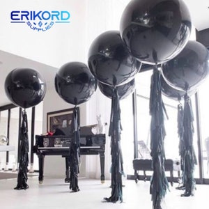 May include: Four large black balloons with black tassel streamers. The balloons are tied together and are floating in the air. The balloons are in front of a window with a white frame. The text "ERIKORD SUPPLIES" is visible in the top left corner of the image.