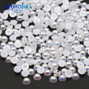 Peut inclure: Un grand nombre de perles rondes irisées blanches avec des dos plats. Les perles sont éparpillées sur une surface grise. ERIKORD SUPPLIES est imprimé en noir sur fond blanc.