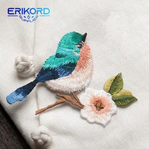 May include: Embroidered patch featuring a blue and green bird perched on a branch with a pink flower. The bird has a white breast and a long tail. The flower has green leaves.