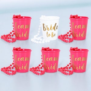 May include: Six pink plastic shot glasses with gold lettering that reads "Team Bride" and white plastic shot glass with gold lettering that reads "Bride to Be". Each shot glass has a pink or white beaded necklace around it.