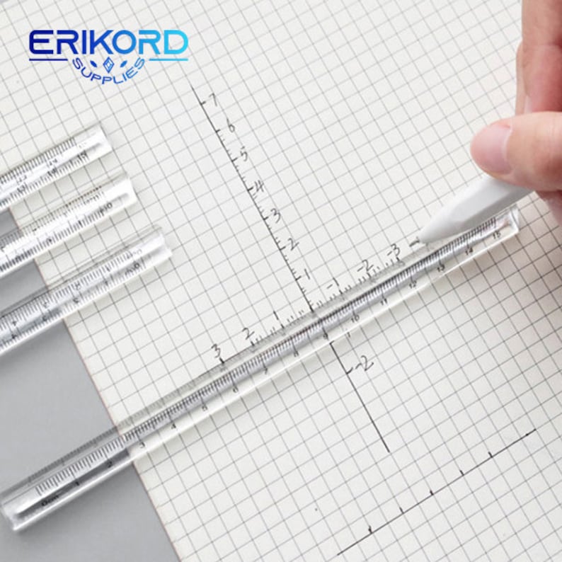 May include: Four clear plastic rulers with inch and centimetre markings on a white gridded paper background. A white pen is lying on top of one of the rulers.