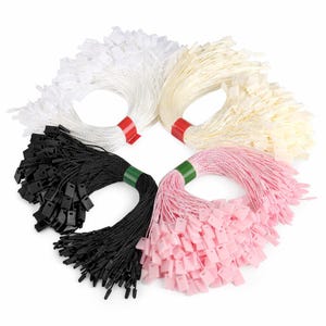 Może przedstawiać: Cztery wiązki etykiet sznurkowych w kolorze białym, kremowym, czarnym i różowym. Każda wiązka jest związana kolorową opaską: czerwoną, zieloną i różową. Etykiety są prostokątne i przymocowane do sznurka.