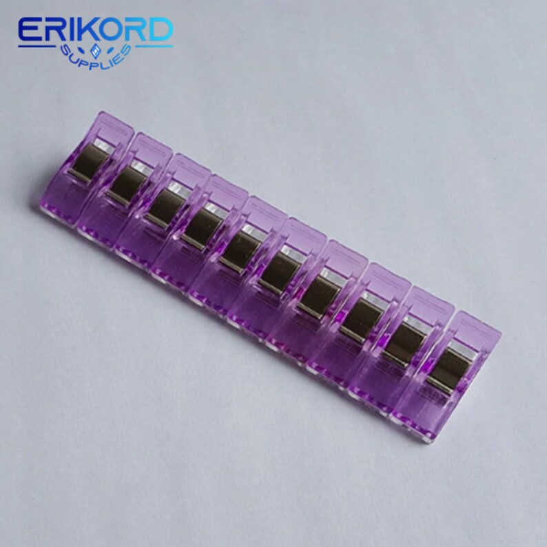 May include: A set of eight purple plastic clips with silver metal spring mechanisms. The clips are arranged in a row on a white surface.