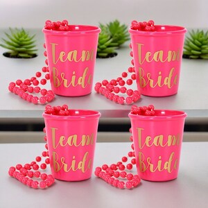 May include: Four pink plastic shot glasses with gold lettering that reads "Team Bride". Each shot glass has a pink beaded necklace wrapped around it.