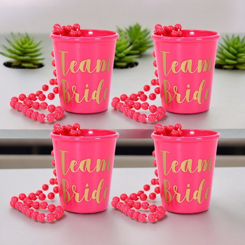 May include: Four pink plastic shot glasses with gold lettering that reads "Team Bride". Each shot glass has a pink beaded necklace wrapped around it.