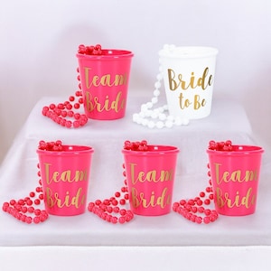 May include: Five pink plastic shot glasses with gold lettering that says "Team Bride" and one white plastic shot glass with gold lettering that says "Bride to Be". Each shot glass has a pink or white beaded necklace around it.