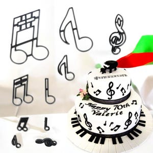 Puede incluir: Un conjunto de 10 plantillas de decoración de pasteles de plástico negro en forma de notas musicales. Las plantillas se muestran sobre una superficie blanca con un pastel de dos niveles decorado con notas musicales negras y un diseño de teclado de piano. El pastel está decorado con el texto "Happy 70th Valerie".