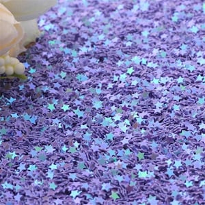 May include: Purple iridescent star-shaped glitter. The glitter is scattered on a surface.