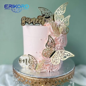 May include: A pink cake decorated with gold glitter butterflies and a gold glitter topper that says "one" and "gunneet".