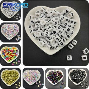 Può includere: Perle di lettere in plastica bianca con numeri e simboli neri. Le perle sono in un piatto a forma di cuore. Ci sono anche altre perle in piatti a forma di cuore separati, tra cui oro, arcobaleno, nero e iridescente.