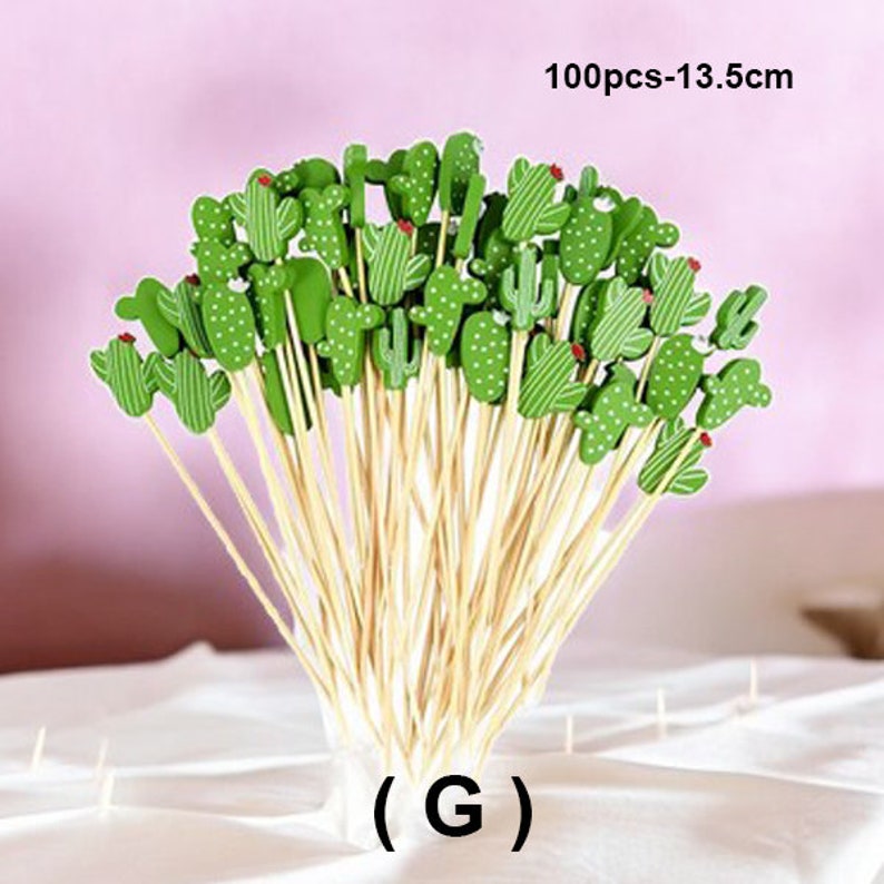 May include: A bunch of 100 green cactus-shaped wooden picks with red accents, each attached to a thin bamboo skewer. The skewers are 13.5 cm long. The picks are arranged in a bouquet shape. The text (G) is visible at the bottom of the image.