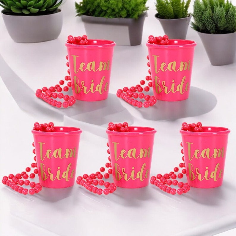 May include: Six pink plastic shot glasses with gold lettering that says "Team Bride" and pink beaded necklaces.