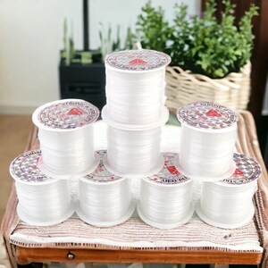 May include: Eight spools of clear elastic string, each wrapped in white paper with red text that reads "Strong & Stretchy 100% Nylon 0.60 mm".