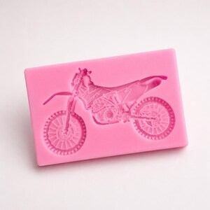 May include: A pink silicone mold featuring a detailed motorcycle design. The mold has a rectangular shape with the motorcycle's outline embossed on the surface, including wheels, engine, and handlebars. Ideal for crafting or baking.