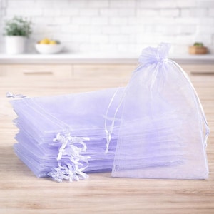 Puede incluir: Un montón de bolsas de organza lavanda transparente con cordón. Las bolsas están hechas de una tela translúcida y están atadas con cintas de satén blancas. Las bolsas están sobre una superficie de madera.