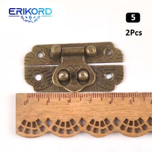 May include: Two antique brass-colored latch clasps with ornate designs. The clasps are closed and have a decorative hinge. The clasps are shown on a wooden ruler with a scalloped edge.