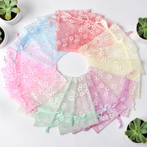 Puede incluir: Una colección de bolsas transparentes con cordón de colores. Cada bolsa está adornada con un delicado estampado floral blanco. Las bolsas son en tonos rosa, azul, amarillo y verde, con lazos de cinta a juego.