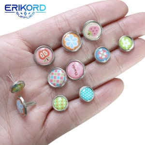 Puede incluir: Surtido de adornos redondos de metal de colores con varios patrones y diseños. Los adornos presentan imágenes de flores, fresas, manzanas, lunares, patrones a cuadros y la palabra "Gracias".