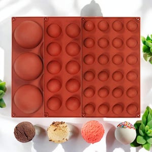 以下が含まれることがあります： 丸いチョコレートキャンディーを作るための3つのセクションを持つ赤いシリコンモールド。モールドには、3つの大きな丸いキャビティと24の小さな丸いキャビティがあります。画像には、完成したチョコレートキャンディーが4つあります。