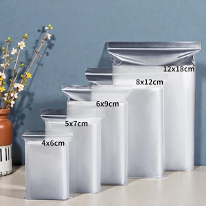 Pode incluir: Um conjunto de cinco sacos de plástico transparentes com fecho zip de tamanhos variados. As dimensões em cm são: 4x6cm, 5x7cm, 6x9cm, 8x12cm e 12x18cm. Os sacos estão dispostos em fila, com o mais pequeno à frente.