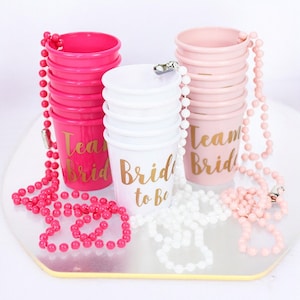 May include: Set of three stacks of plastic cups with beaded necklaces. The cups are pink, white, and pink. The pink cups have gold lettering that says "Team Bride" and "Team Bride". The white cup has gold lettering that says "Bride to Be".