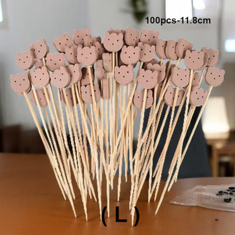 May include: A bunch of 100 brown flower-shaped picks with wooden sticks, 11.8 inches long. The picks are arranged in a fan shape, with the sticks pointing downwards. The text "100pcs-11.8cm" is visible in the top right corner of the image. The text "(L)" is visible in the bottom center of the image.