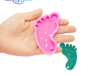 May include: Pink silicone mold shaped like a baby foot. A teal glitter baby foot is shown next to the mold.