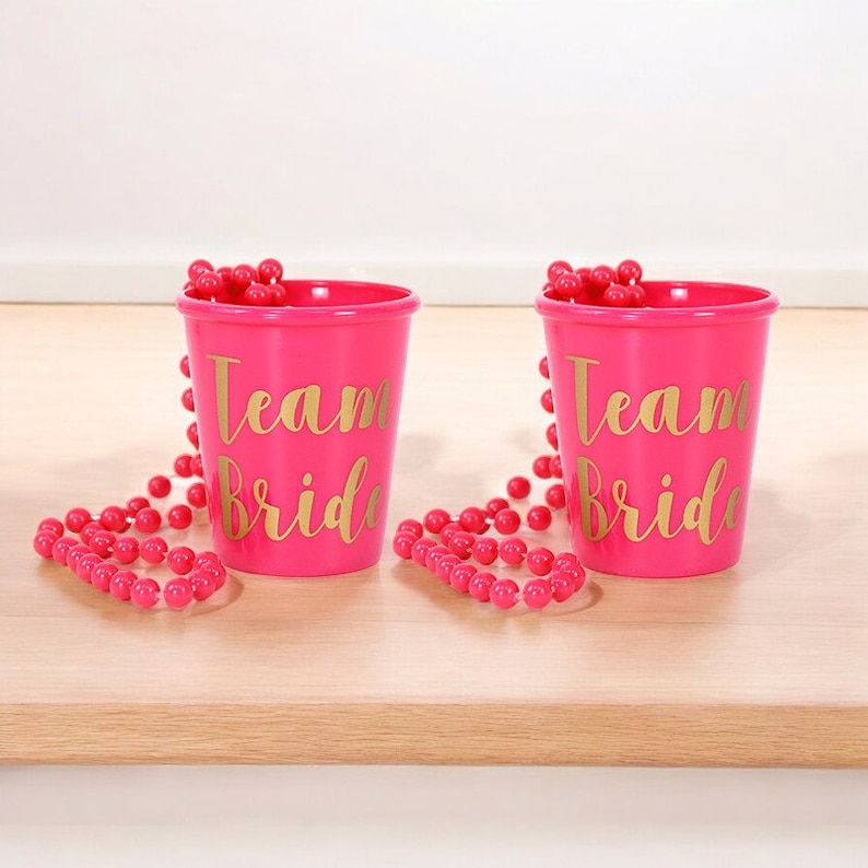 May include: Two pink plastic cups with gold lettering that says "Team Bride". Each cup has a pink beaded necklace wrapped around it.