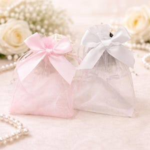 以下が含まれることがあります： ピンクと白の、サテンのリボンで結ばれた2つのシアーオーガンジーのギフトバッグ。バッグには未知のアイテムが詰められ、パールをあしらった柔らかいフローラルの背景に配置されています。
