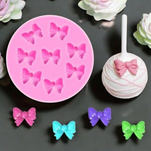May include: Pink silicone mold with nine bow-shaped cavities.  A cake pop with a pink bow on top is shown next to the mold.  Five small bows in different colors are shown on a gray surface.
