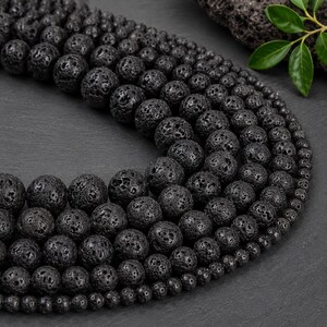 May include: A collection of spherical black lava beads arranged in a curved formation. The beads feature a porous, textured surface with varying diameters, ranging from larger beads to smaller ones, creating a gradient of sizes in the display.