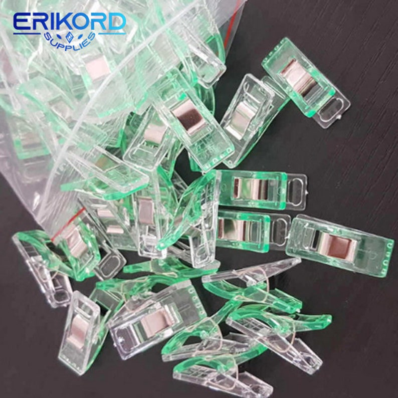 May include: A pile of clear plastic sewing clips with metal spring closures. The clips are green tinted and are used for holding fabric together while sewing.