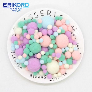 May include: A white plate filled with a variety of colourful pom poms. The pom poms are in shades of pink, green, blue, and purple. The plate has text around the edge, including the words "RICCUTI SAVOIE" and "GALETTE".