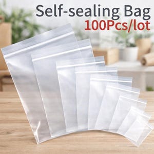 Puede incluir: Bolsas de plástico transparentes y autosellantes de varios tamaños, con cierre de cremallera, dispuestas sobre una superficie de madera. La inscripción "Self-sealing Bag 100Pcs/lot" es visible. Las bolsas son transparentes.