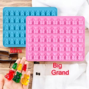 Könnte beinhalten: Zwei Silikonformen für Gummibärchen, eine blaue und eine rosa. Die blaue Form ist mit "Mini" und die rosa Form mit "Big Grand" beschriftet. Neben den Formen befinden sich Gummibärchen in verschiedenen Farben.