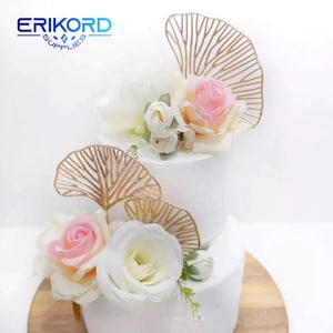 Könnte beinhalten: Zweistöckiger weißer Kuchen, verziert mit rosa und weißen Rosen und goldenen, blattartigen Kuchenaufsätzen. Der Kuchen steht auf einem Holzkuchenständer.