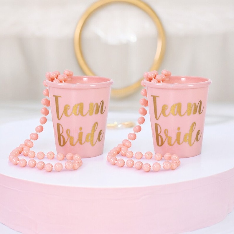 May include: Two pink plastic cups with gold lettering that says "Team Bride" and pink beaded necklaces attached to the cups.