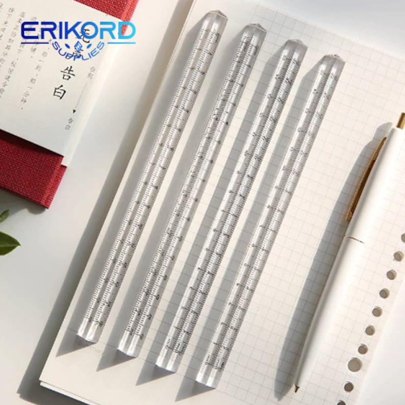 May include: Four clear plastic rulers with black markings, each measuring 76 cm long, are laid out on a white gridded surface. A white pen with a gold accent is also visible.