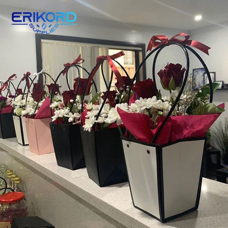 Puede incluir: Cinco arreglos florales en cajas de regalo blancas y negras con rosas rojas y flores blancas. Las cajas tienen un asa negra y un fondo blanco. Las cajas est&aacute;n sobre un mostrador blanco.