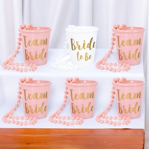 May include: Six pink plastic cups with gold lettering that says "Team Bride" and one white plastic cup with gold lettering that says "Bride to Be". Each cup has a pink or white beaded necklace around it.