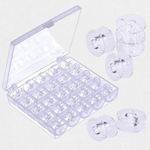 Puede incluir: Una caja de almacenamiento transparente para bobinas de máquina de coser con tapa abierta, llena de bobinas transparentes. Varias bobinas sueltas están esparcidas alrededor de la caja.