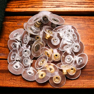 Puede incluir: Un montón de respaldos de pendientes de plástico transparente con postes de metal. Algunos postes son plateados, otros son dorados y algunos son una combinación de ambos.