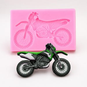 May include: A green, black, and white toy motorcycle with a detailed design, positioned in front of a pink silicone mold of a motorcycle. The toy motorcycle has black tires and a detailed engine.