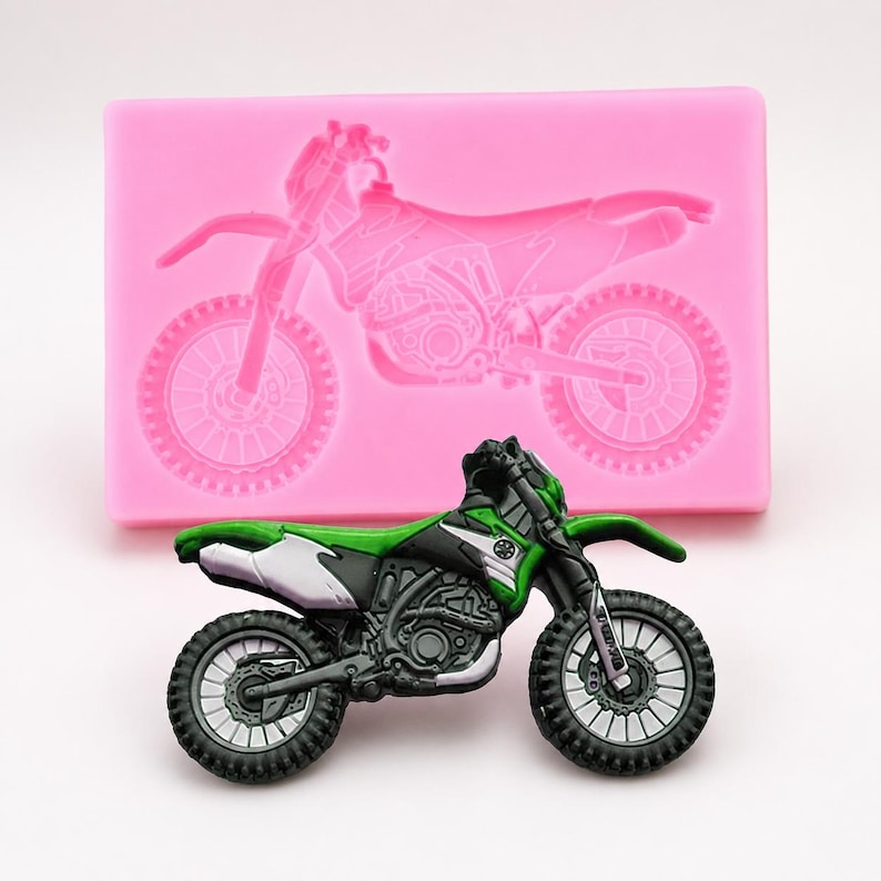 May include: A green, black, and white toy motorcycle with a detailed design, positioned in front of a pink silicone mold of a motorcycle. The toy motorcycle has black tires and a detailed engine.