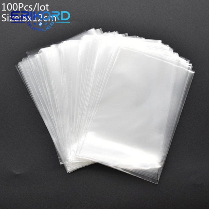 Op de afbeelding: Een stapel van 100 doorzichtige plastic zakjes, elk met een afmeting van 20,3 x 30,5 cm. De zakjes zijn gevouwen en gestapeld op een zwart oppervlak.