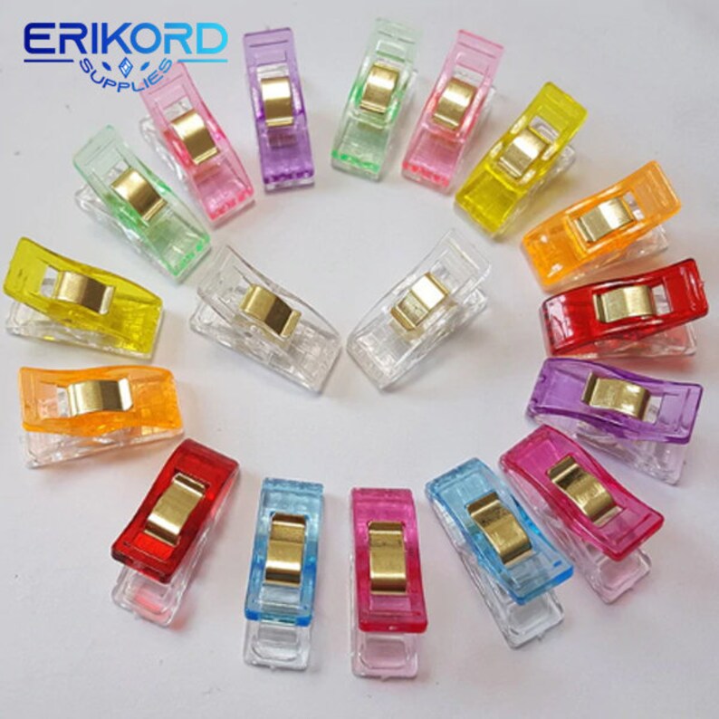 May include: A set of 15 colorful plastic clips for sewing and crafting. The clips are arranged in a circle on a white background. The clips are clear, with a variety of colors, including red, orange, yellow, green, blue, purple, and pink.