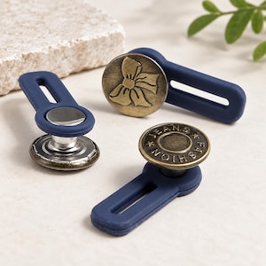 May include: Three adjustable waist extenders featuring dark blue silicone tabs. One extender has a brass-toned button with a bow design, another has a plain silver-toned button, and the third has a brass-toned button embossed with the text JEANS FASHION.
