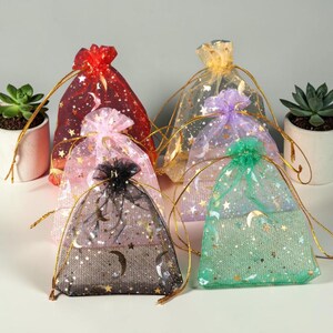 May include: Six colorful organza gift bags with gold drawstrings. The bags are in shades of red, pink, black, yellow, purple, and green. Each bag is decorated with gold stars and crescent moons. The bags are ideal for small gifts or party favors.
