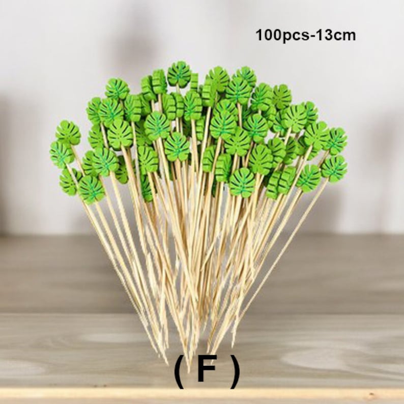 May include: A bunch of 100 green leaf picks on bamboo skewers, each measuring 13 cm long. The leaves are arranged in a bouquet shape, with the skewers pointing upwards. The text "100pcs-13cm" is visible in the top right corner of the image. The text "(F)" is visible in the bottom center of the image.