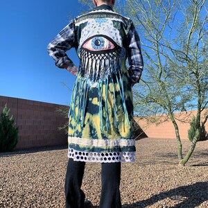 May include: A long denim vest with a tie-dye pattern in shades of blue and yellow. The vest has a large eye applique with blue and white floral details and fringe trim.